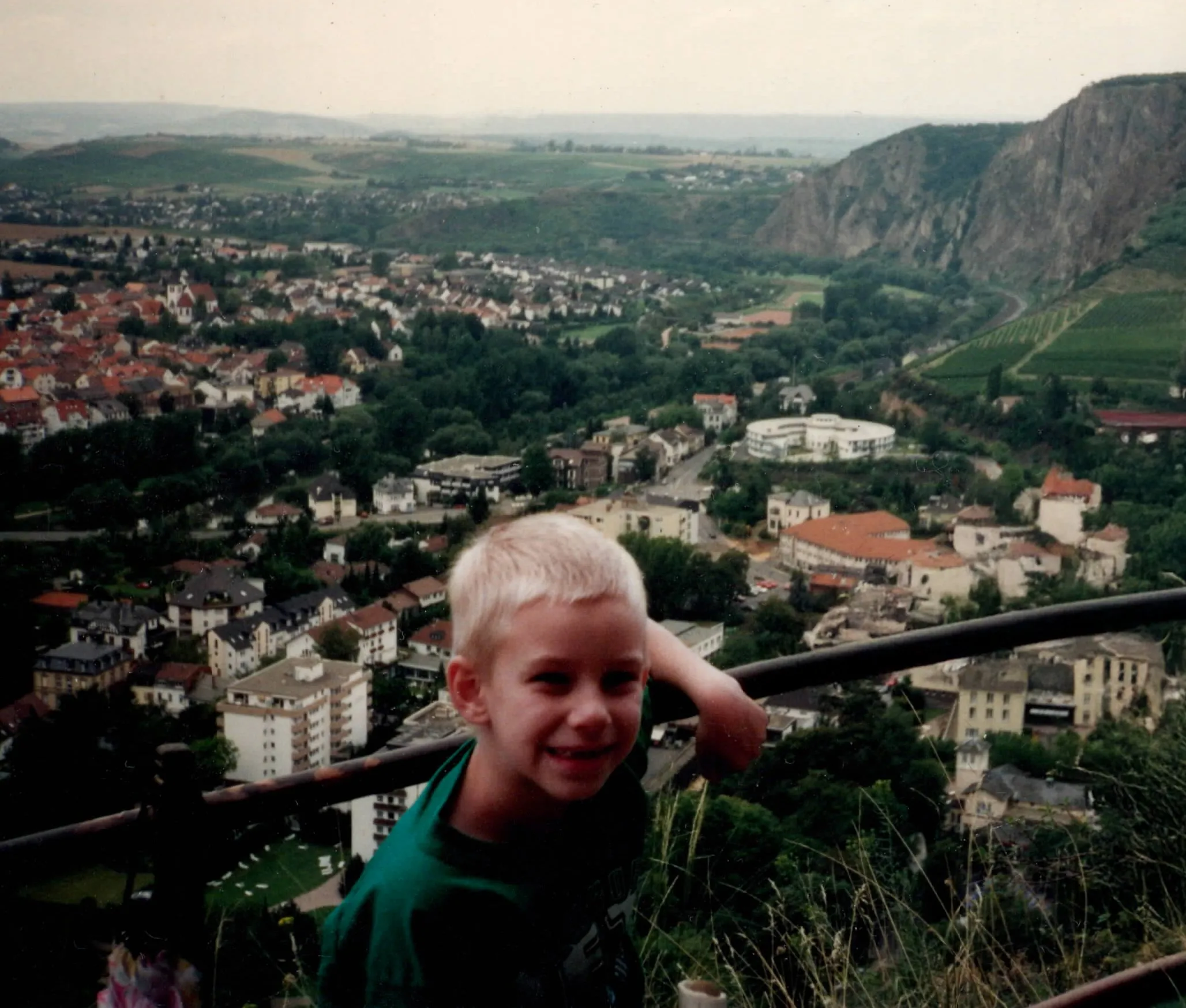 A young Erik, smiling and giving a thumbs-up gesture, with a German cityscape and green hills behind him.