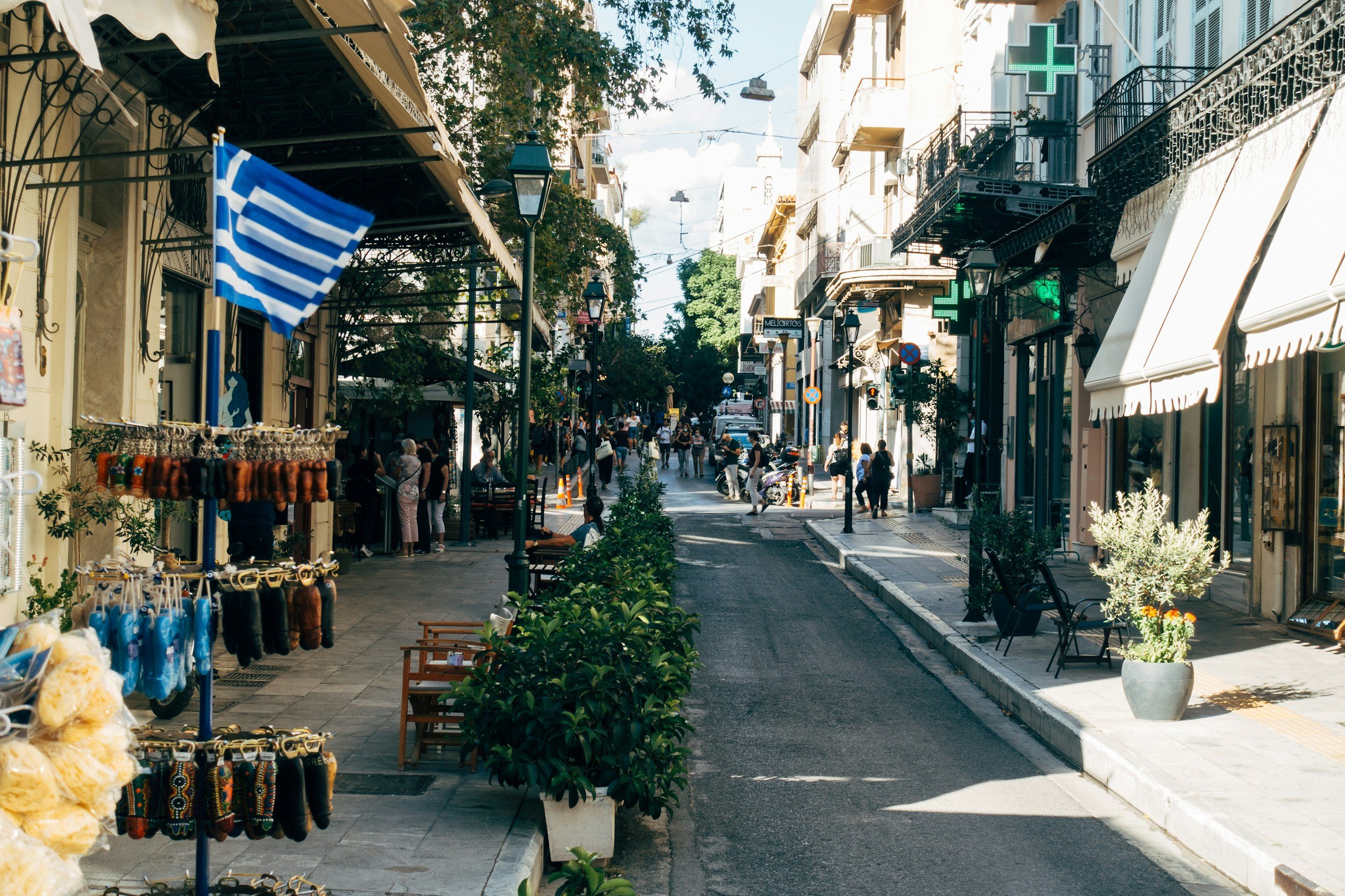 A shaded Athens street with shopfronts, plants and a Greek flag